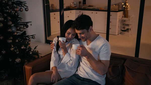 Feliz hombre abrazando novia con taza de té cerca del árbol de Navidad en la sala de estar - foto de stock