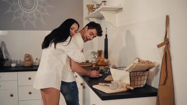 Happy young woman in white shirt hugging cheerful boyfriend in kitchen — Stock Photo