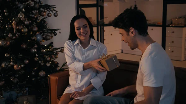 Happy woman presenting gift to joyful boyfriend near christmas tree — Stock Photo