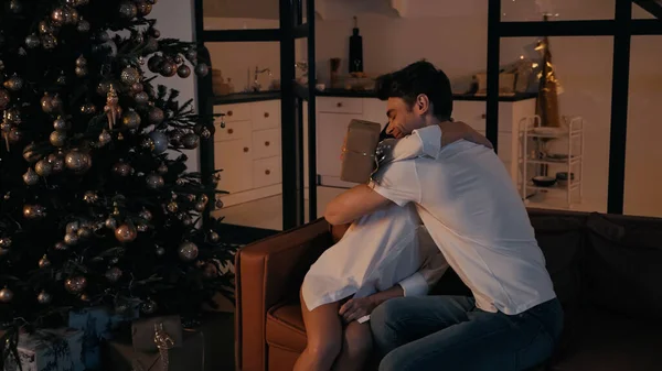 Hombre alegre abrazando a la mujer mientras está sentado en el sofá cerca del árbol de Navidad con regalos - foto de stock