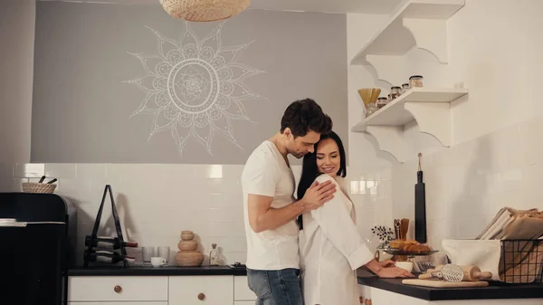 Hombre feliz coqueteando y abrazando a mujer joven en camisa blanca en la cocina moderna - foto de stock