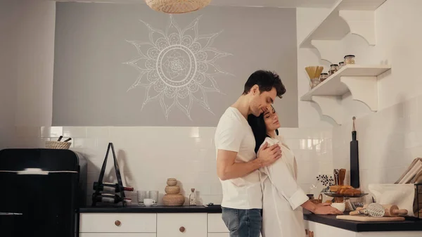 Homem feliz abraçando jovem mulher em camisa branca na cozinha moderna — Fotografia de Stock