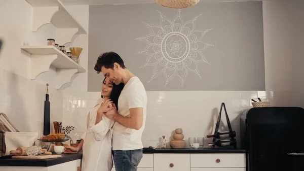 Homme heureux étreignant jeune femme brune en chemise blanche dans la cuisine moderne — Photo de stock