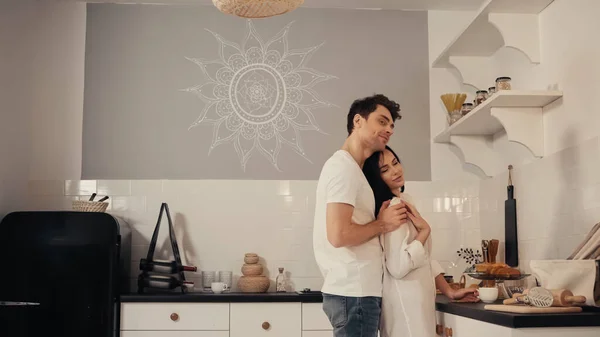 Hombre complacido abrazando a mujer joven y sonriente en camisa blanca en cocina moderna - foto de stock