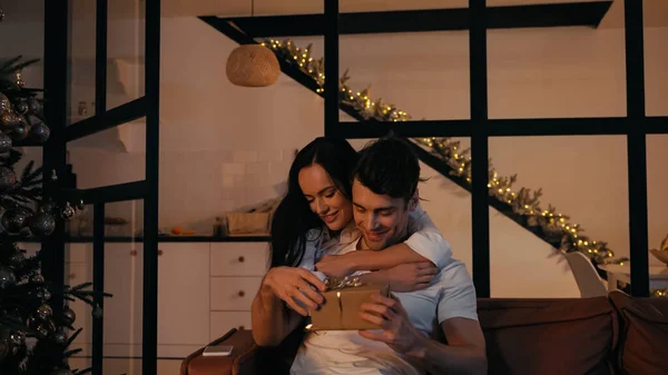 Mulher alegre ajudando presente aberto de namorado sentado no sofá perto de smartphone e árvore de Natal — Fotografia de Stock