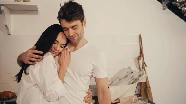 Morena mujer joven en camisa blanca abrazo complacido novio en cocina moderna - foto de stock