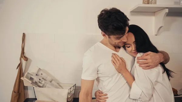 Morena complacida joven en camisa blanca abrazándose con el novio en la cocina moderna - foto de stock