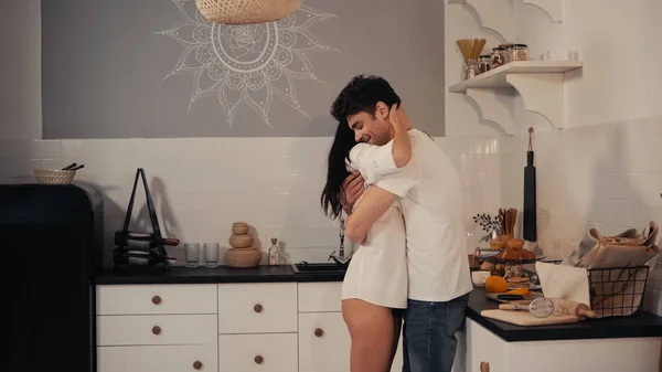 Morena mujer joven en camisa blanca abrazando novio positivo en cocina moderna - foto de stock