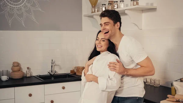 Happy man embracing cheerful girlfriend and laughing in kitchen — Stock Photo
