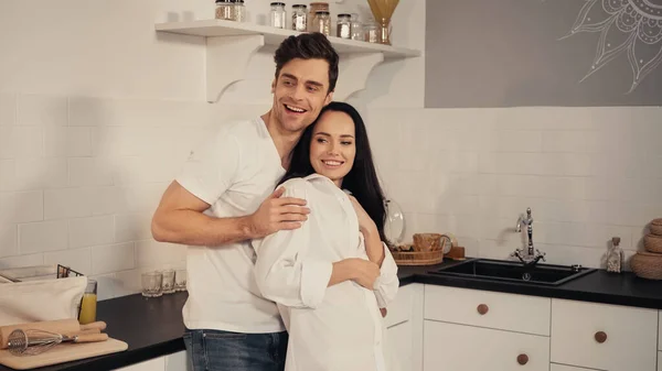 Happy man embracing cheerful girlfriend and smiling in kitchen — Stock Photo