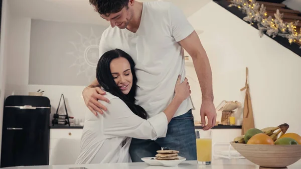 Mujer alegre abrazando al hombre cerca de panqueques y jugo de naranja en la cocina con decoración de Navidad - foto de stock