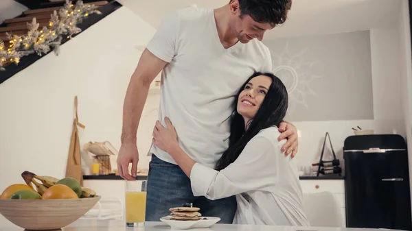 Счастливая женщина обнимает мужчину рядом с блинами и апельсиновым соком на кухонном столе — стоковое фото