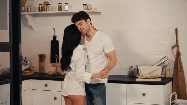 Улыбающийся мужчина обнимает женщину в рубашке во время танцев на современной кухне — стоковое фото