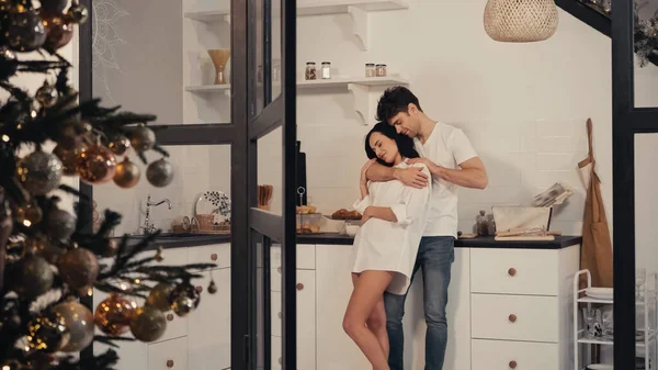 Щаслива молода пара обіймається на кухні з прикрашеною ялинкою за дверима — стокове фото