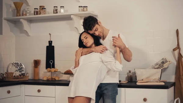 Мужчина обнимает брюнетку и чувственную женщину в белой рубашке, стоя на современной кухне — стоковое фото