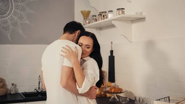 Uomo abbraccio bruna e sensuale donna in camicia bianca in cucina — Foto stock