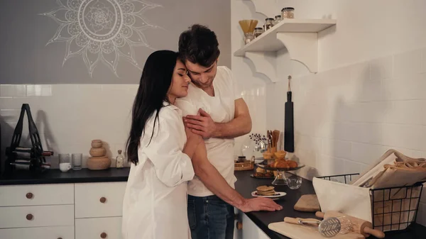 Brünette und sinnliche Frau in weißem Hemd umarmt glücklichen Mann in moderner Küche — Stockfoto