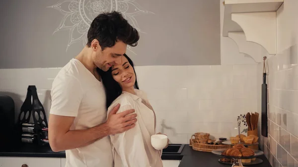 Hombre feliz abrazando morena y mujer sonriente en camisa blanca de pie con taza de café - foto de stock