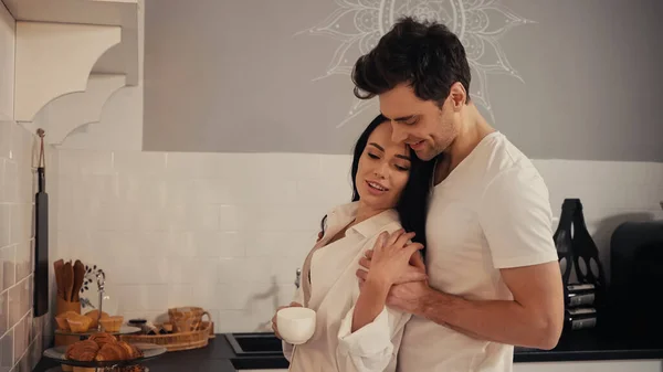 Hombre feliz abrazando morena y sensual mujer en camisa blanca de pie con taza de café - foto de stock