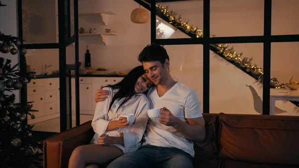 Happy woman holding cup and leaning on shoulder of boyfriend while sitting on sofa near decorated christmas tree — Stock Photo