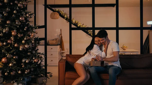 Feliz mujer abrazando alegre novio apertura presente cerca decorado árbol de Navidad - foto de stock