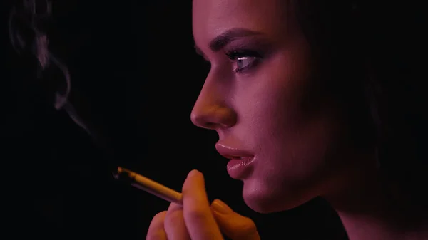 Вид збоку молодої жінки, що тримає розмиту сигарету на чорному тлі — стокове фото