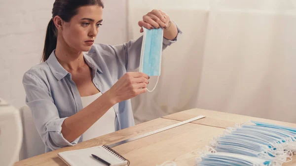 Brunette seamstress checking medical mask near ruler and notebook in atelier — Stock Photo