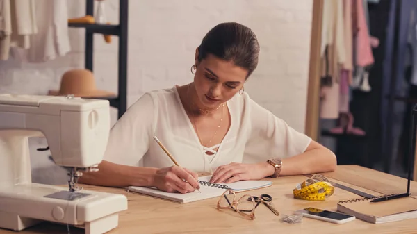 Diseñador morena escribiendo en el portátil cerca de la máquina de coser y el teléfono inteligente en el taller - foto de stock