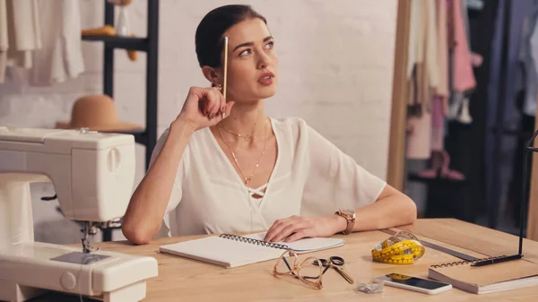 Pensive designer sitting near notebook and sewing machine in atelier — Stock Photo