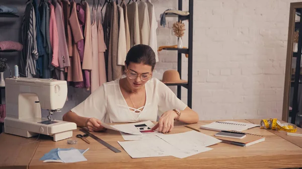 Seamstress holding sketches near sewing machine and notebooks in atelier — Stock Photo