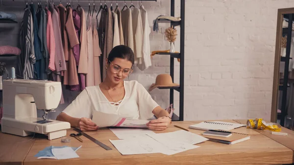 Diseñador en anteojos mirando bocetos cerca de máquina de coser y cuadernos en atelier - foto de stock