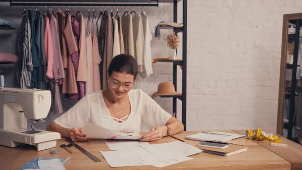 Smiling designer holding sketches near smartphone and sewing machine in atelier — Stock Photo