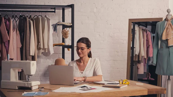 Designer em óculos usando laptop perto de esboços e máquina de costura no atelier — Fotografia de Stock
