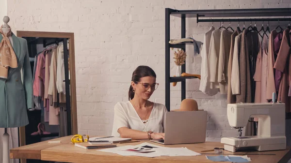 Designer sorrindo usando laptop perto da máquina de costura e esboços no atelier — Fotografia de Stock