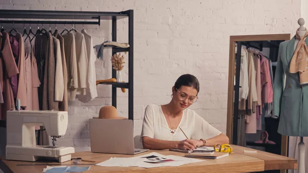 Bruna designer scrittura su notebook vicino a dispositivi e macchina da cucire in atelier — Foto stock