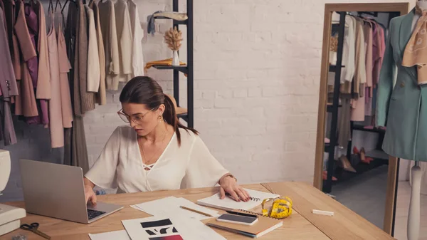 Seamstress using laptop near notebooks and sketches in workshop — Stock Photo