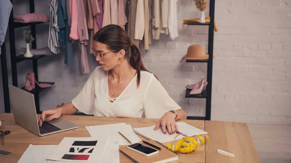 Morena costurera usando portátil cerca de cuadernos y bocetos en atelier - foto de stock