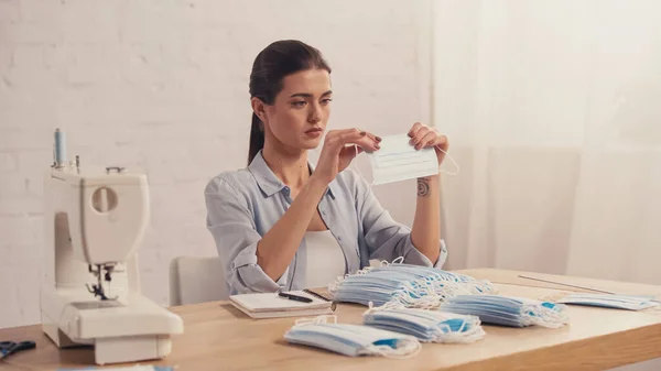 Brunette seamstress holding medical mask near sewing machine in atelier — Stock Photo