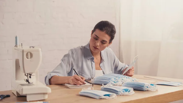 Costurera sosteniendo máscara médica y escritura en el cuaderno cerca de la máquina de coser - foto de stock