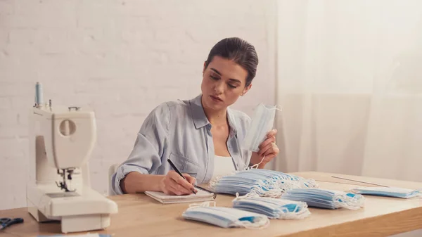 Seamstress writing on notebook near medical masks and sewing machine in atelier — Stock Photo