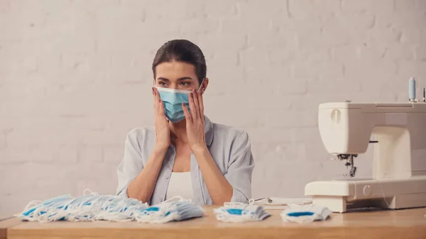 Brunette seamstress in protective mask sitting near sewing machine in workshop — Stock Photo