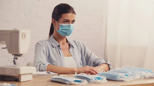 Brünette Näherin in Schutzmaske sitzt neben Nähmaschine im Atelier — Stockfoto