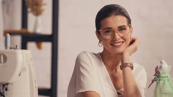 Lächelnde Näherin mit Brille blickt in Kamera neben verschwommener Schaufensterpuppe und Nähmaschine im Atelier — Stockfoto