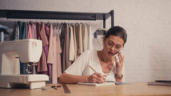 Seamstress talking on smartphone and writing on notebook near sewing machine in atelier — Stock Photo