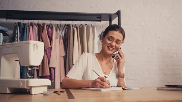 Costurera positiva hablando en el teléfono inteligente y escribiendo en el portátil cerca de tijeras y máquina de coser - foto de stock