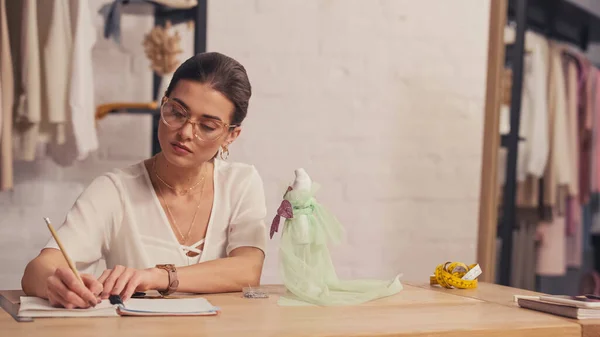 Seamstress writing on notebook near mannequin and tape measure on table — Stock Photo