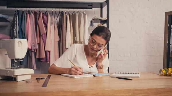 Seamstress falando no celular e escrevendo no notebook perto da máquina de costura no atelier — Fotografia de Stock