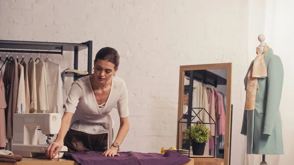 Diseñador tocando tela cerca de la máquina de coser y maniquí en atelier - foto de stock