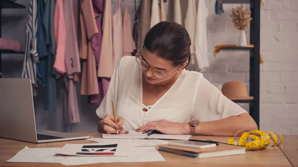 Diseñador escribiendo en un cuaderno cerca de dispositivos y bocetos en el taller - foto de stock