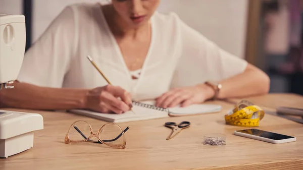 Vista cortada de óculos perto da máquina de costura e desenhista borrado escrevendo no notebook no atelier — Fotografia de Stock